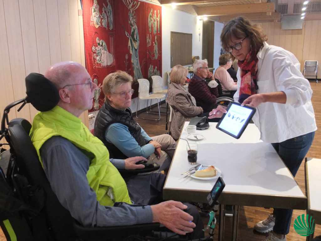 Frau Zang zeigt einem Herrn im Rollstuhl eine App am Tablet